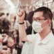 Man standing in the righthand-side of the frame in focus wearing a medical mask holding the strap on a subway with other riders behind him out of focus.