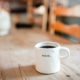 White coffee mug, full of coffee, sitting on long wooden table with wooden chairs out of focus in the background