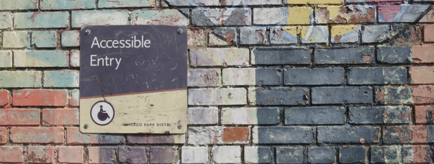Close-up of a faded multicolor mural on a brick wall with a sign to the left that says, "Accessible Entry" with a small wheelchair accessible icon